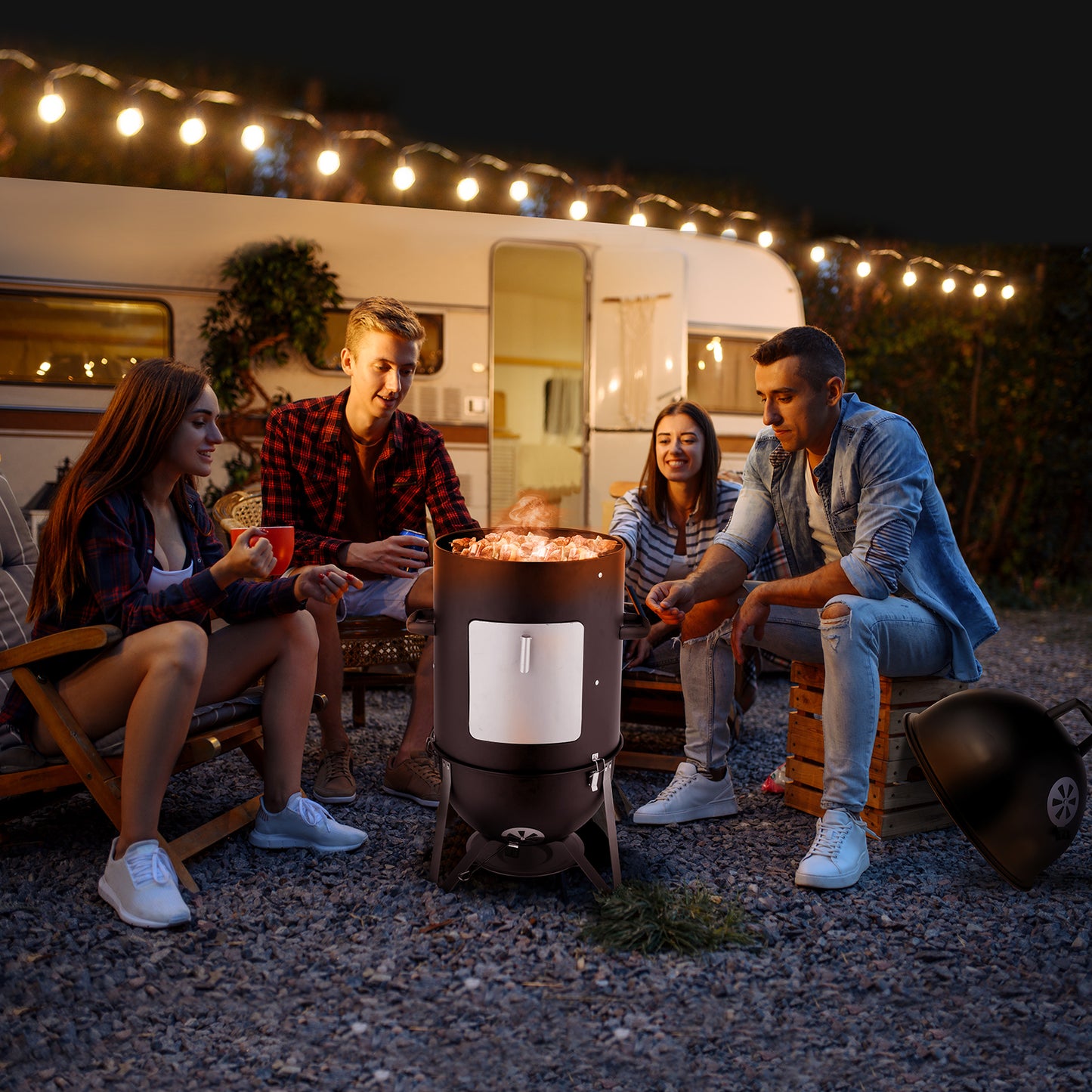Four people sitting around a portable fire pit at night near a camper trailer with string lights.