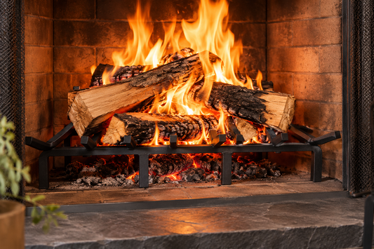 A close-up of a black metal fireplace log rack holding cut firewood with bright orange and yellow flames burning above, set inside a brick fireplace with a dark stone hearth in the foreground.